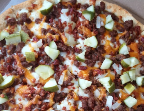 A close-up of a pizza topped with ground meat, bacon, onions, and green pickles on a golden crust.