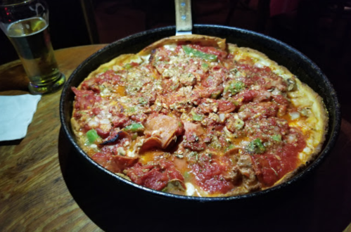 A deep-dish pizza topped with pepperoni, green peppers, and a chunky tomato sauce, served on a wooden table.