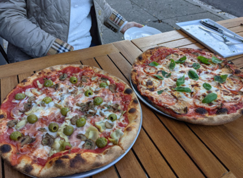 Two pizzas on a wooden table: one topped with green olives, ham, and artichokes; the other with cheese and basil.