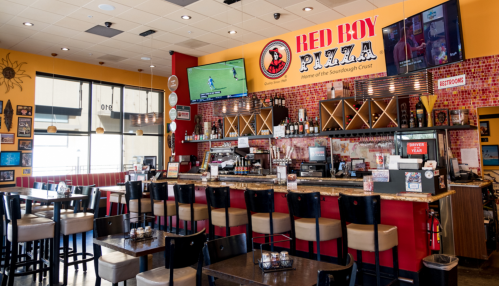 Interior of Red Boy Pizza, featuring a bar area, tables, and TVs displaying sports, with colorful decor.