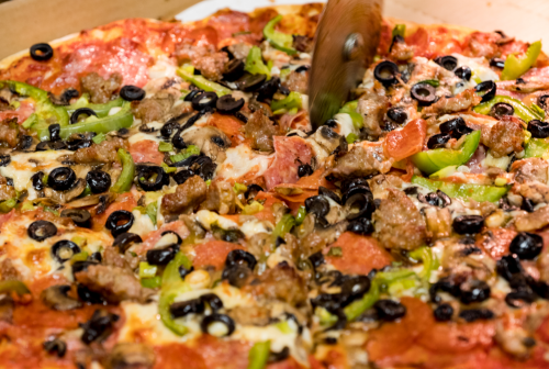 A close-up of a pizza topped with pepperoni, green peppers, black olives, and sausage, being sliced with a pizza cutter.