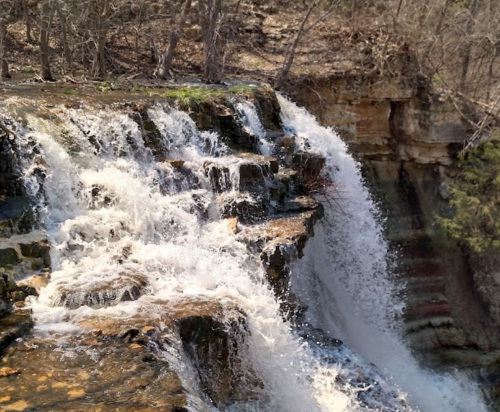A cascading waterfall flows over rocky cliffs, surrounded by bare trees and earthy tones.