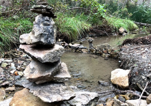 A stack of rocks beside a small stream, surrounded by greenery and pebbles.