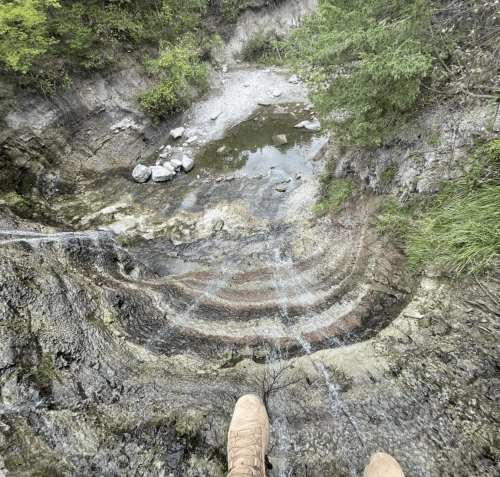 A person's feet in boots at the edge of a rocky cliff overlooking a small stream and lush greenery below.