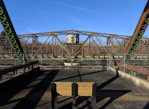 A weathered metal bridge with a control tower, viewed from a platform with a wooden bench in the foreground.