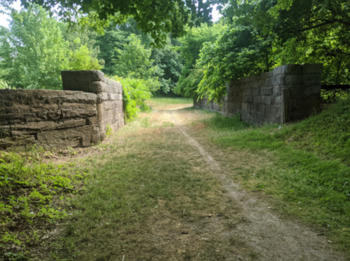 A grassy path leads through stone walls, surrounded by lush greenery and trees.