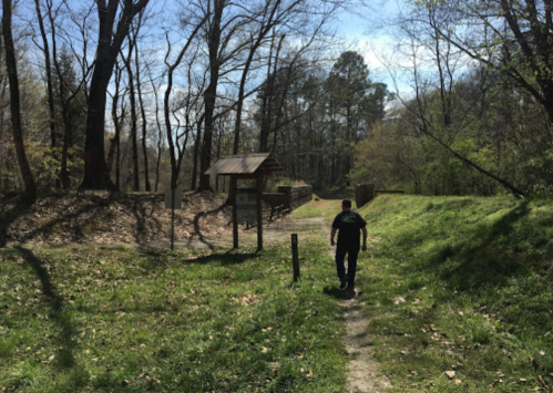 A person walking on a dirt path through a wooded area, with trees and a signpost in the background.
