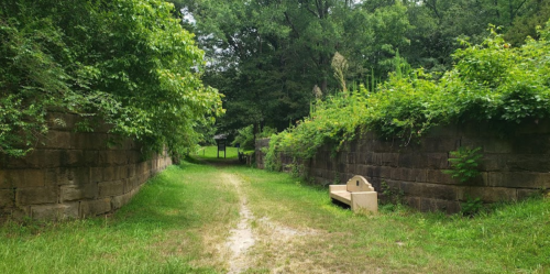 A grassy path flanked by stone walls, with lush greenery and a bench on one side, leading into a wooded area.