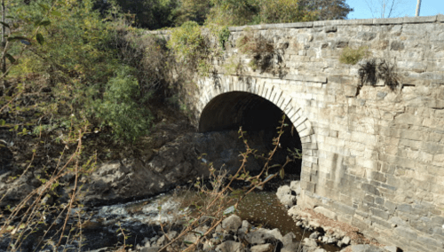 A stone arch bridge over a small river, surrounded by greenery and rocky terrain.