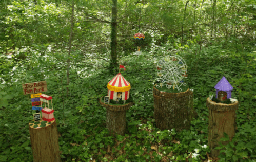 Miniature carnival scene in a forest, featuring a circus tent, ferris wheel, and colorful attractions on tree stumps.