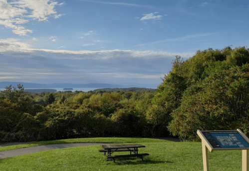 A scenic view of a lush landscape with a picnic table and informational sign, overlooking a body of water and distant hills.