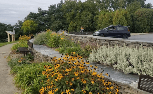 A landscaped area with flowers and greenery, a stone wall, and a parked car in a scenic outdoor setting.