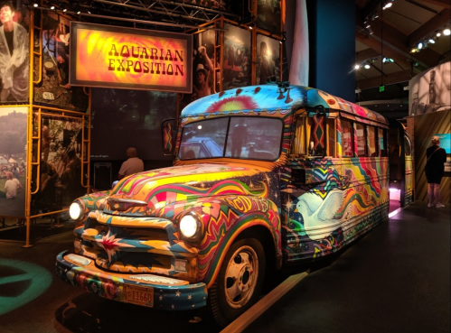 A colorful, psychedelic bus on display at an exhibition, surrounded by vibrant artwork and historical photos.