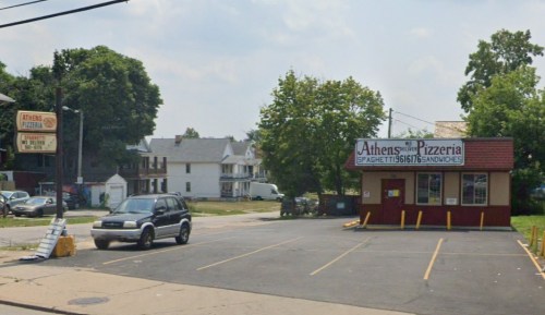 A pizzeria with a sign reading "Athens Pizzeria" and a parking lot, surrounded by trees and residential buildings.