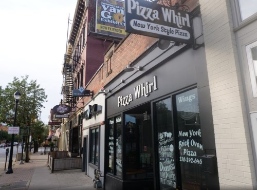 Exterior of Pizza Whirl, a New York style pizza restaurant, with signage and nearby buildings on a city street.