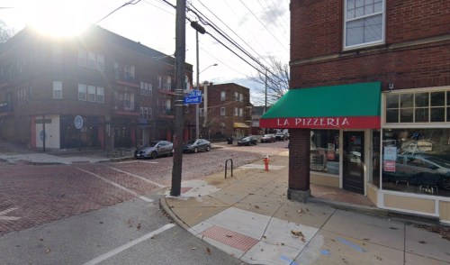 A street view of a corner with a pizzeria, brick buildings, and parked cars under a partly cloudy sky.