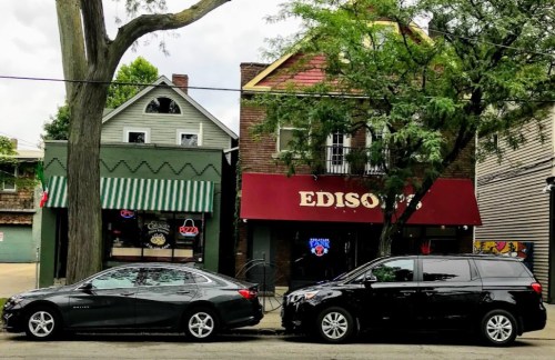 Two parked cars in front of a brick building with a red awning labeled "EDISON'S" and a green-striped storefront.