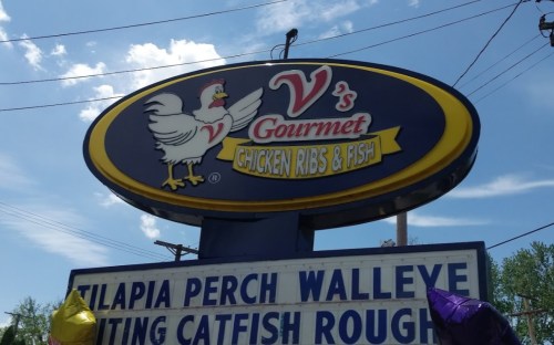 Sign for V's Gourmet Chicken Ribs & Fish, featuring a chicken graphic and menu items like tilapia and walleye.