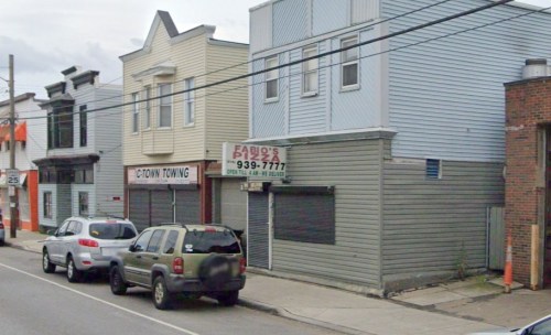 A street view of a pizza shop and a towing service, with parked cars along the road.