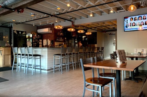 A modern restaurant interior featuring a bar, wooden tables, and a TV displaying news. Warm lighting creates a cozy atmosphere.