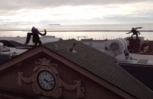 Two ornate statues on a rooftop overlook a calm sea with an island in the distance, under a cloudy sky.