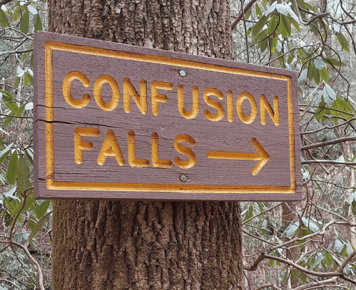Wooden sign reading "Confusion Falls" with an arrow pointing to the right, mounted on a tree in a forested area.