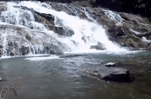 A cascading waterfall flows over rocky terrain into a serene pool below, surrounded by lush greenery.