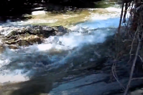 A flowing river with rocky banks, surrounded by trees and sunlight reflecting on the water's surface.