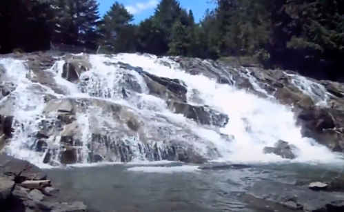 A cascading waterfall flows over rocky terrain, surrounded by lush green trees under a clear blue sky.