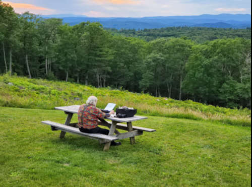 An elderly person sits at a picnic table, working on a laptop with a scenic mountain view in the background.