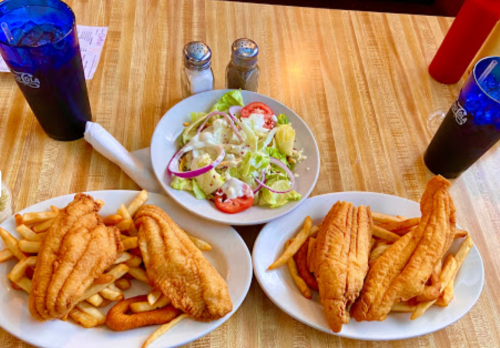 Two plates of fried fish and fries, a salad with tomatoes and onions, and two blue drinks on a wooden table.