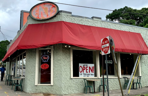 A restaurant with a red awning and "OPEN" sign, featuring outdoor seating and a textured gray exterior.