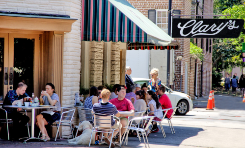 Outdoor café scene with people dining at tables, a dog resting nearby, and a sign for "Clary's" in the background.
