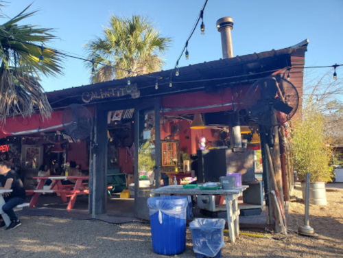 A rustic outdoor dining area with picnic tables, palm trees, and a colorful building under a clear blue sky.