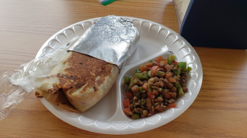 A wrapped burrito on a paper plate, accompanied by a side of diced vegetables and lentils.