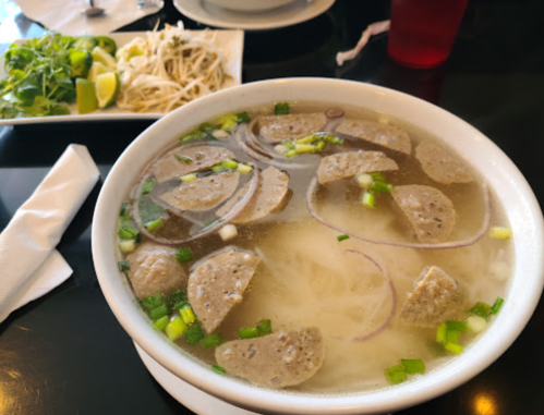 A bowl of soup with meatballs, noodles, and green onions, accompanied by a plate of herbs and lime.