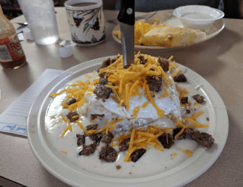 A plate of breakfast featuring eggs, sausage, cheese, and gravy, topped with a knife, with a side of a burrito and coffee.