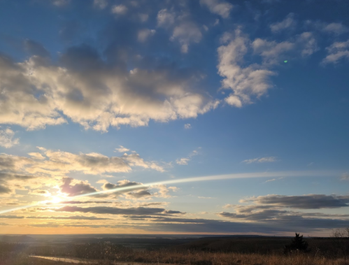 A scenic view of a sunset with clouds and a horizon, casting warm light over a grassy landscape.