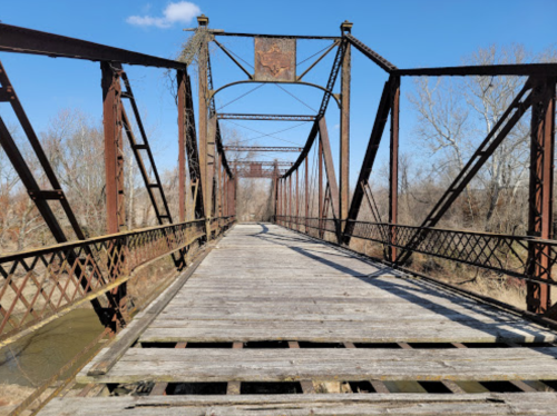 A weathered metal bridge with wooden planks, spanning a river under a clear blue sky.
