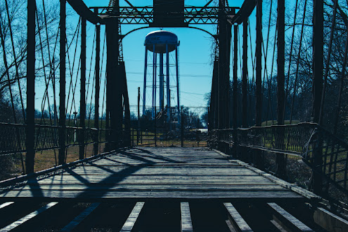 A weathered wooden bridge leads to a water tower in the distance, framed by bare trees and a clear blue sky.