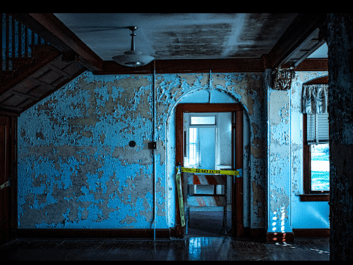 An abandoned room with peeling blue paint, a doorway, and caution tape across the entrance. Light filters in through windows.