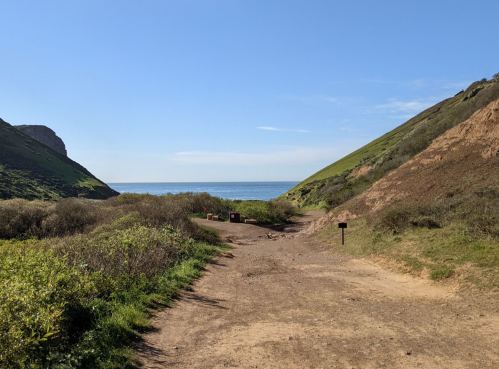 A dirt path leads to the ocean, flanked by green hills and shrubs under a clear blue sky.