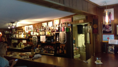 A cozy bar with a wooden counter, shelves lined with various bottles, and patrons enjoying drinks in a warm atmosphere.