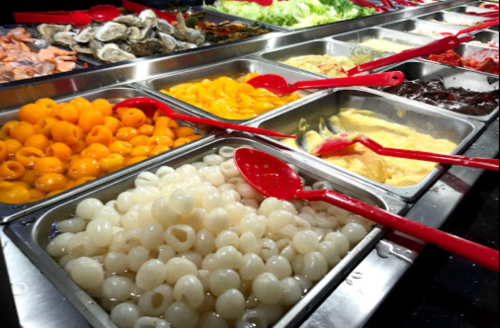 A variety of colorful food items in metal trays, with red serving spoons, displayed at a buffet.