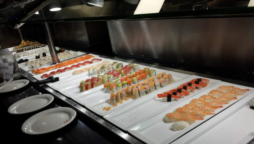 A variety of sushi rolls and sashimi displayed on white trays at a sushi bar, with empty plates in the foreground.