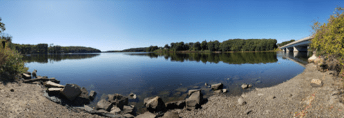 A serene river scene with clear blue skies, calm water, and a bridge in the background, surrounded by greenery.