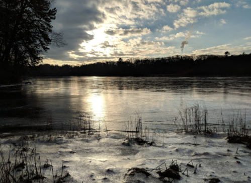 A serene winter landscape featuring a frozen lake, reflecting clouds and sunlight, surrounded by trees.