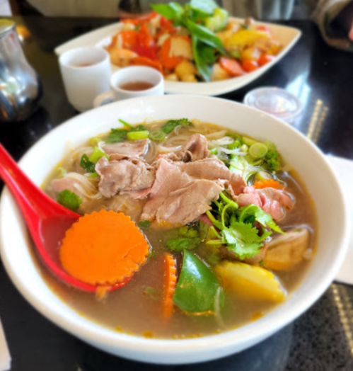 A bowl of noodle soup with meat, herbs, and vegetables, alongside a plate of colorful stir-fried vegetables.
