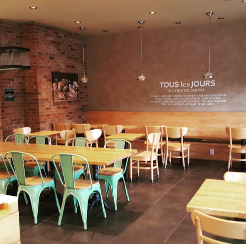 A cozy bakery interior with wooden tables, green chairs, and a brick wall featuring the name "Tous les Jours."