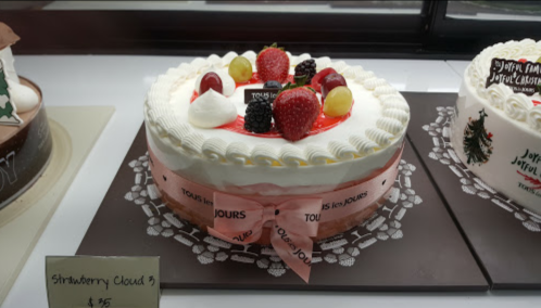A decorated strawberry cake topped with berries and cream, displayed in a bakery case with a pink ribbon.